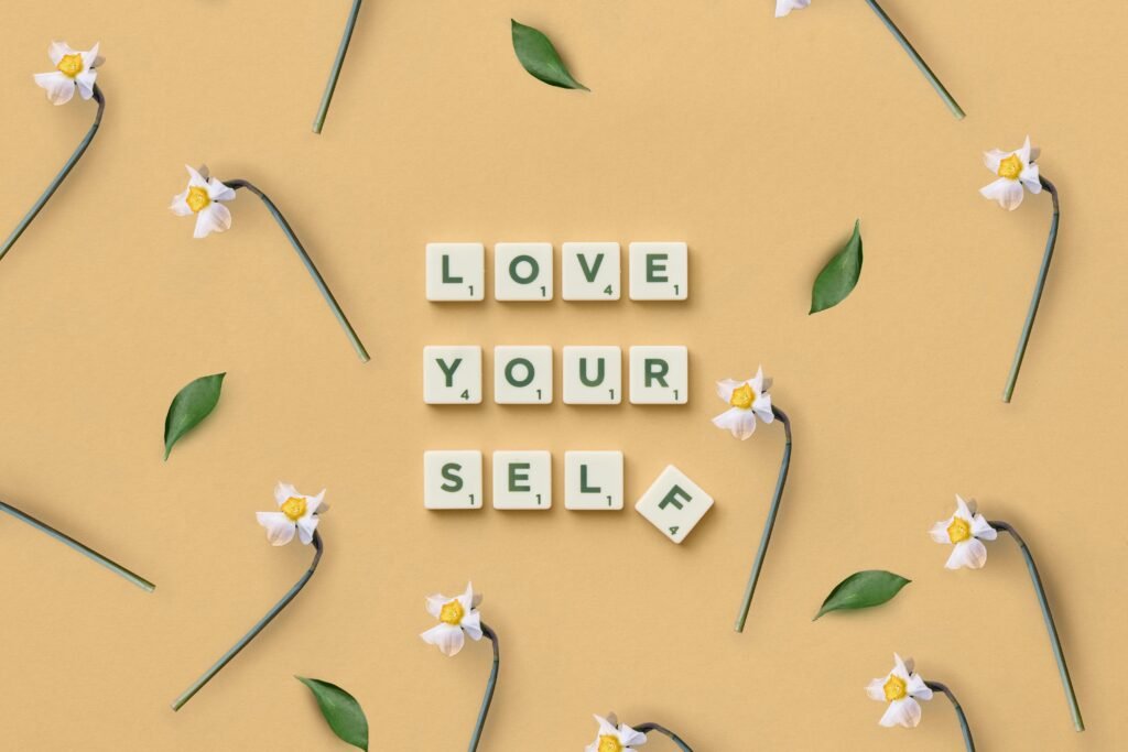 Scrabble tiles spelling 'Love Yourself' surrounded by flowers and leaves on a flat surface.